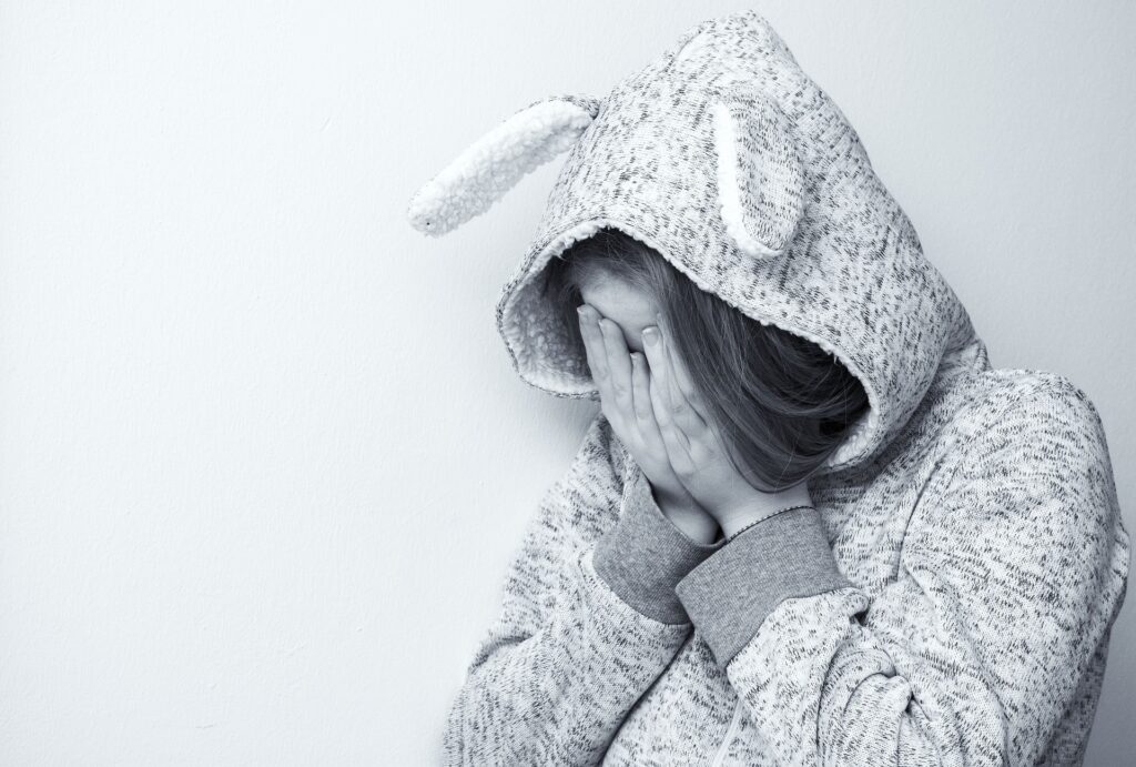 Person sitting with their face covered by their hands, wearing a hooded top, representing excessive apologising, anxiety, shame, low self-esteem, and fear of upsetting others.